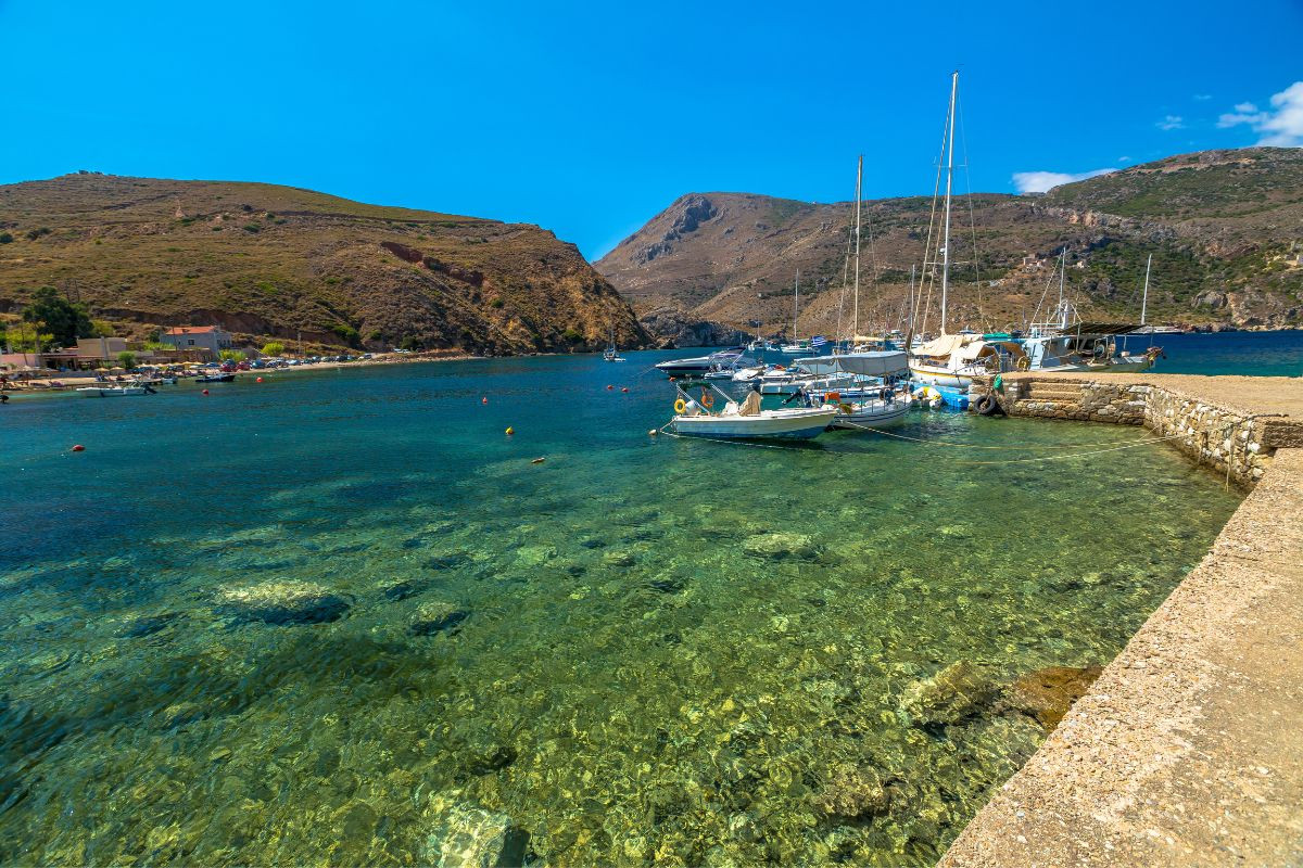 Boats are docked at a clear, calm bay surrounded by rocky hills in the Peloponnese region, with a small village visible on the shoreline under a bright blue sky.